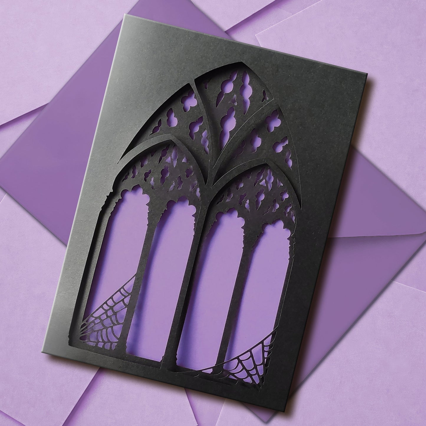 A handmade black greeting card with two intricate papercut layers of a gothic cathedral window with spiderwebs in the corners. Your choice of blood red or lilac purple shows between the window arches. Comes with a matching colour envelope.