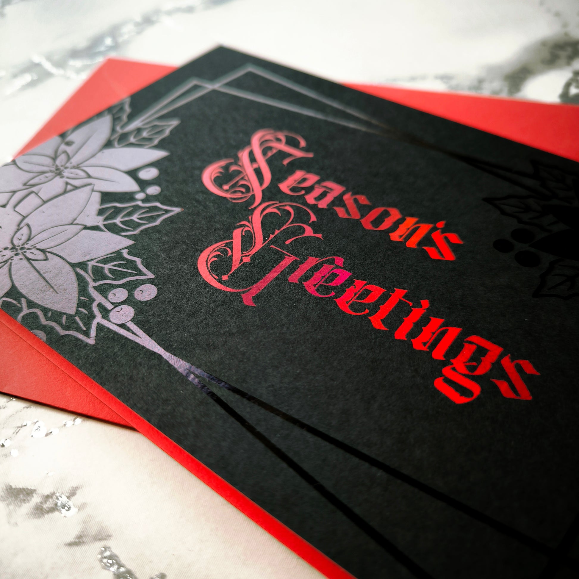 A5 landscape handmade Christmas card on black with red foil “Season’s Greetings” lettering and black foil poinsettia and holly motifs in the corners. Subtle border lines. Includes red envelope. Blank inside.