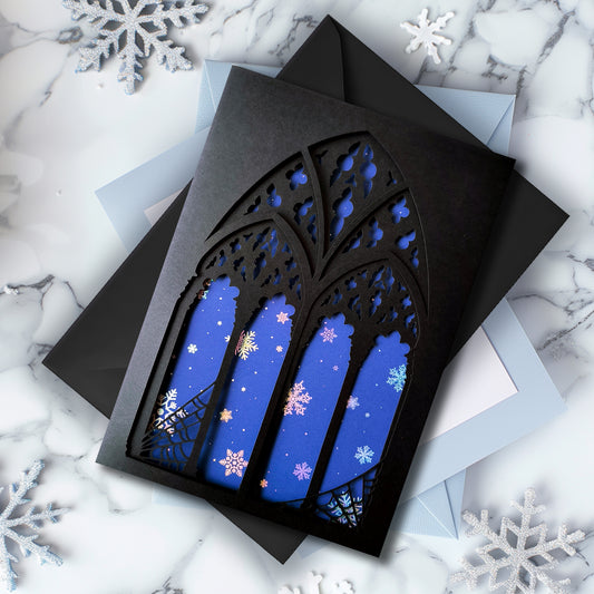 A5 handmade layered papercut Christmas card with black gothic cathedral arch window cutout and spiderweb corners, midnight blue background with holographic snowflakes. Portrait format. Includes black envelope. Blank inside.