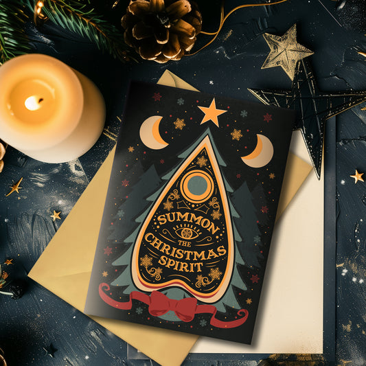 A5 glossy portrait Christmas card on black with ornate Ouija planchette reading “Summon the Christmas Spirit”, moons, stars, snowflakes and pine tree shapes with gold and green details. Cream envelope. Inside has pale green border with snowflake corners, blank area.