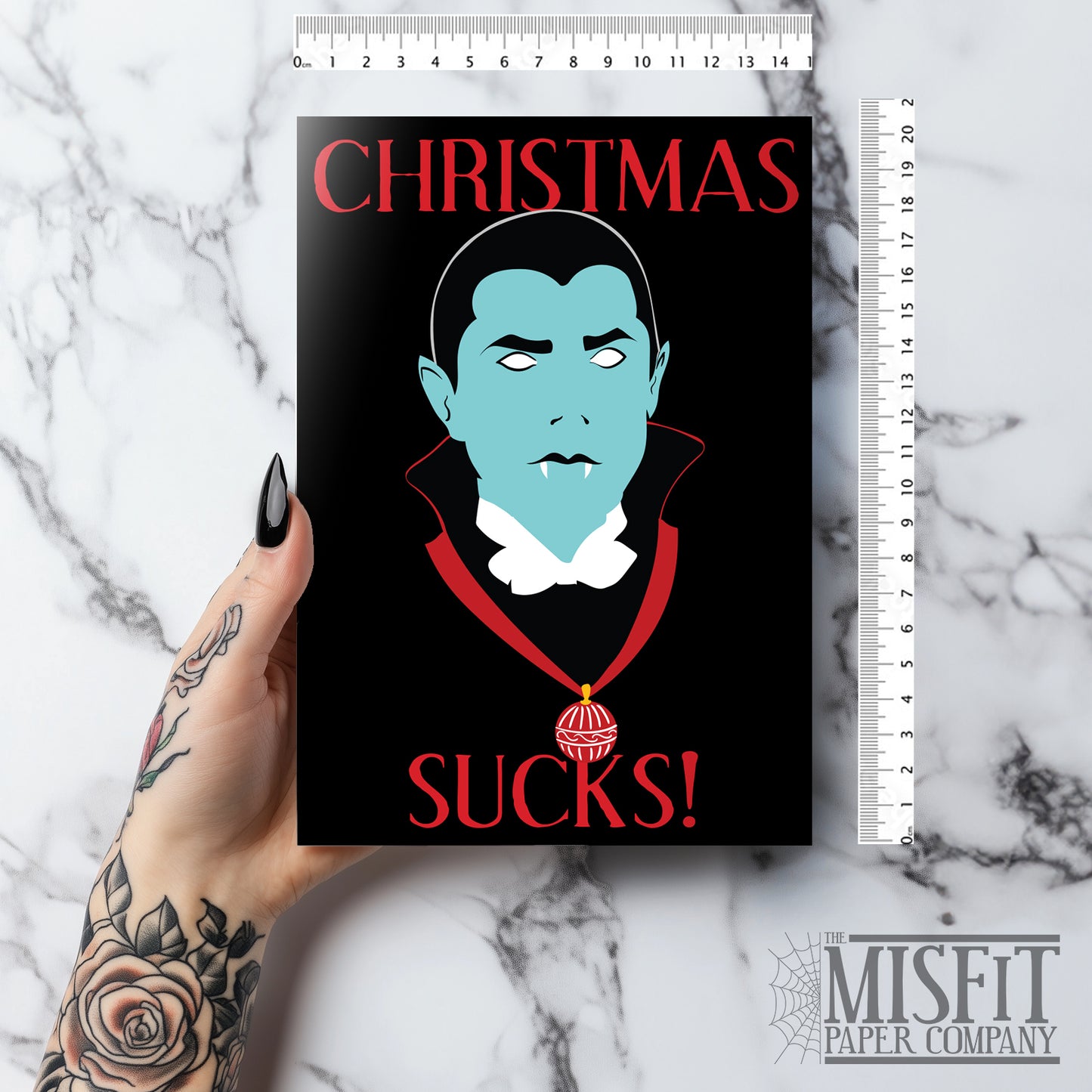 A5 glossy portrait greeting card with graphic vampire on black background, pale blue face, white eyes and fangs, red collar and bauble. Red serif text reads “CHRISTMAS SUCKS!”. Gothic, spooky style. Includes red envelope. Blank inside.