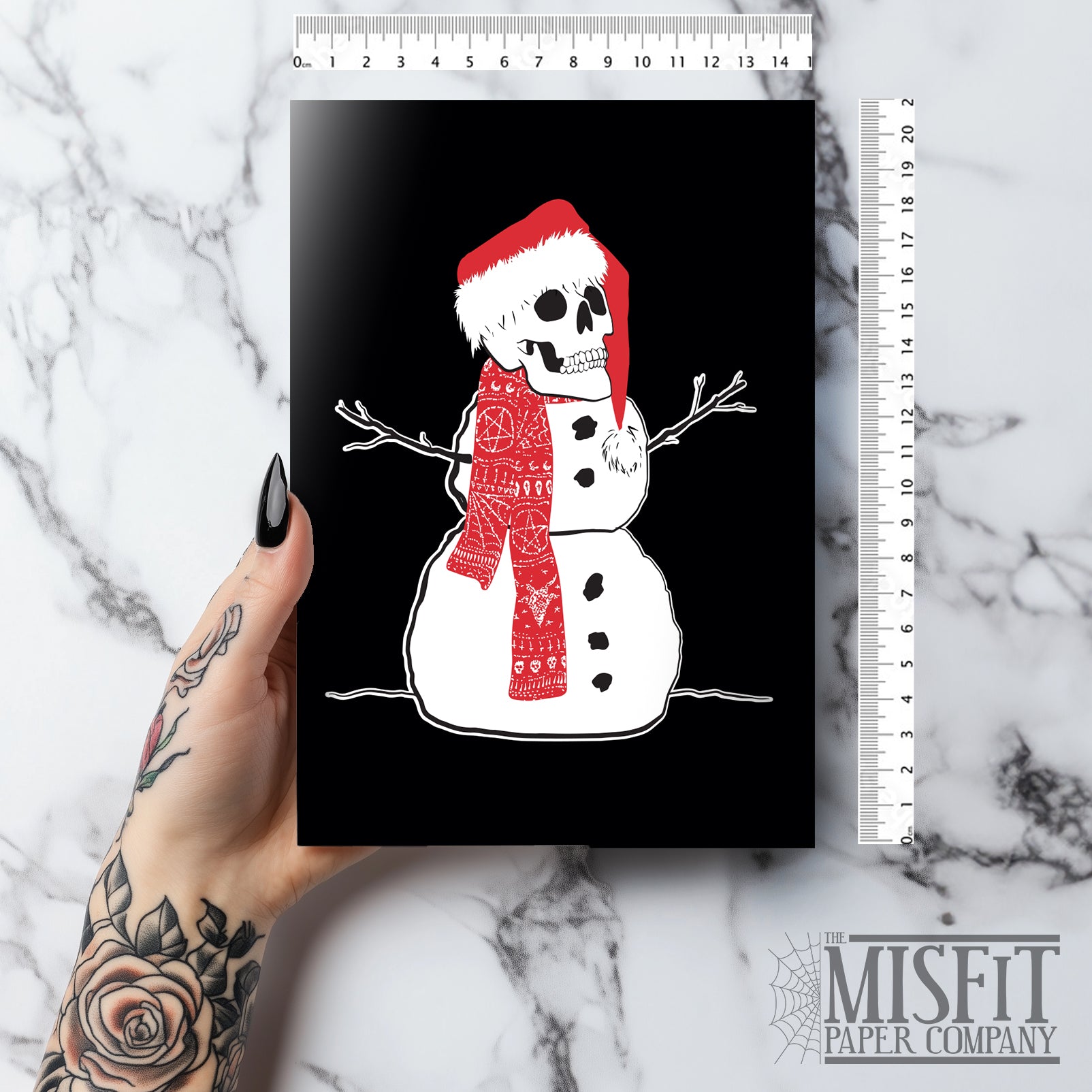 A5 glossy portrait Christmas card with white snowman on black background, skull head wearing a Santa hat and patterned red scarf, stick arms and coal buttons. Clean graphic style. Includes red envelope. Blank inside.