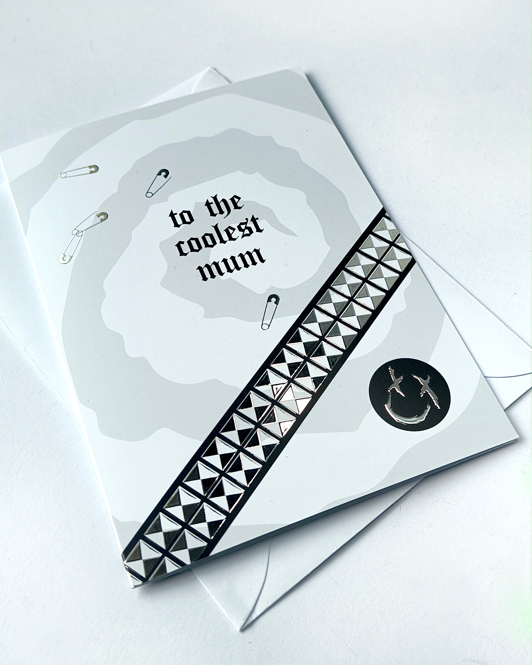 A5 greeting card reading “to the coolest mum” with gothic emo styling, featuring chrome stud belt graphic, safety pin details and a black smiley badge, paired with a white envelope.