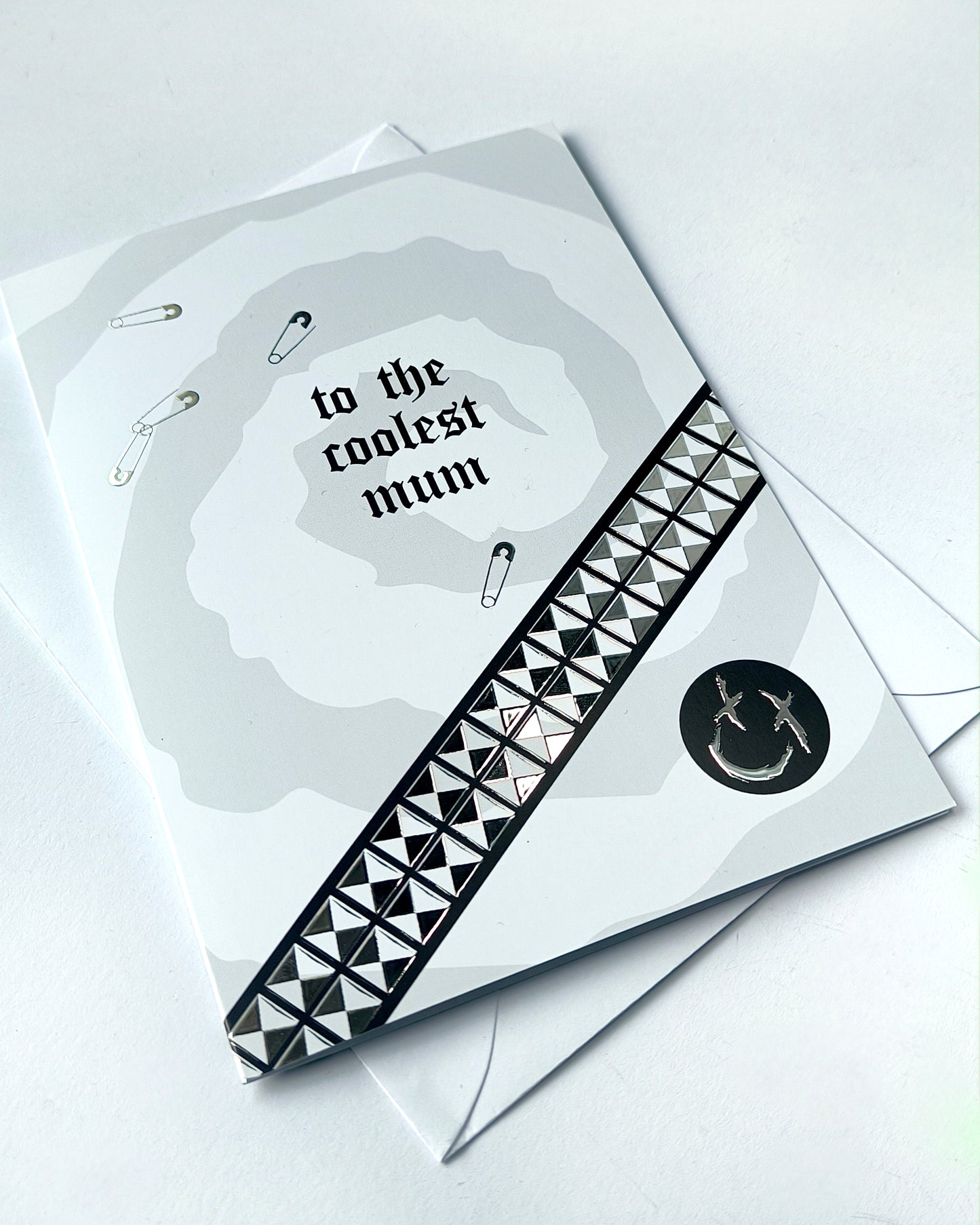 A5 greeting card reading “to the coolest mum” with gothic emo styling, featuring chrome stud belt graphic, safety pin details and a black smiley badge, paired with a white envelope.