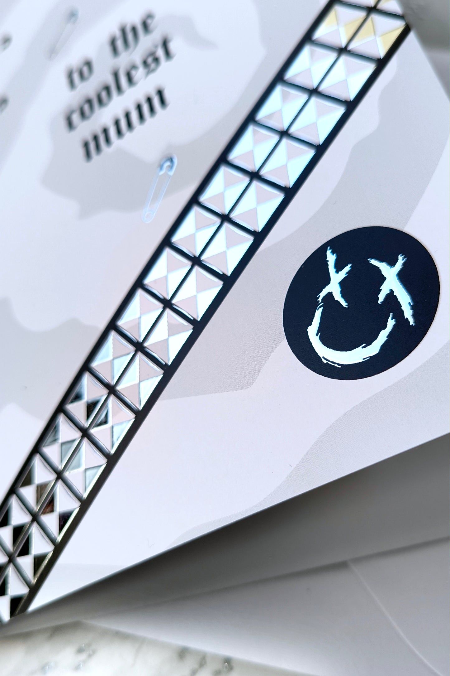 A5 greeting card reading “to the coolest mum” with gothic emo styling, featuring chrome stud belt graphic, safety pin details and a black smiley badge, paired with a white envelope.
