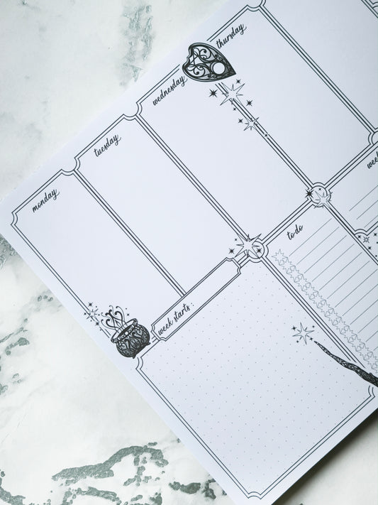 Undated weekly planner pad with black and white line-art frames and doodles. Blank boxes for each day, notes area, colouring-friendly layout. Desk pad style on smooth paper.
