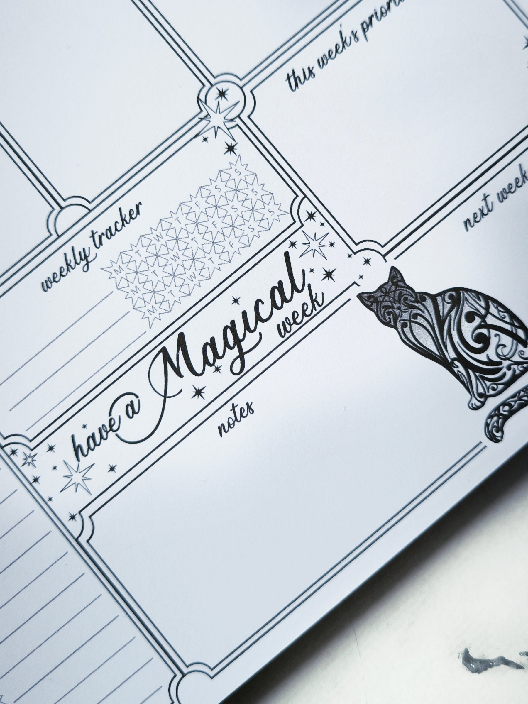 Undated weekly planner pad with black and white line-art frames and doodles. Blank boxes for each day, notes area, colouring-friendly layout. Desk pad style on smooth paper.