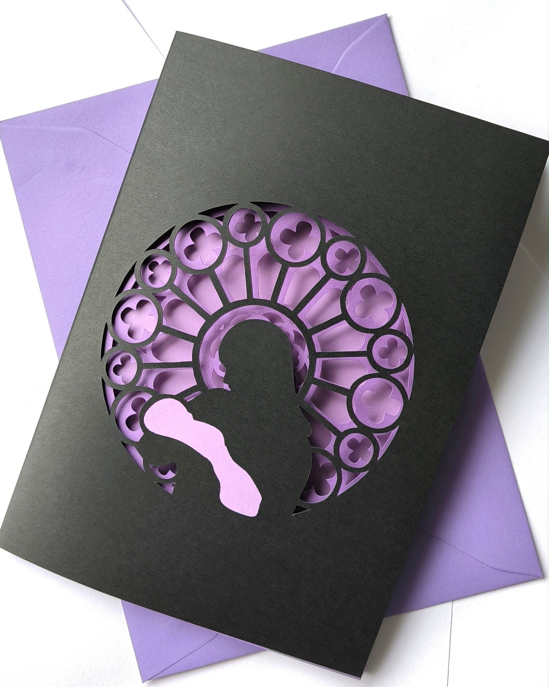 Handmade papercut greeting card, with a layered rose window in purple and black. Features a mother and baby silhouette in front of the gothic cathedral window design. Includes matching purple envelope.