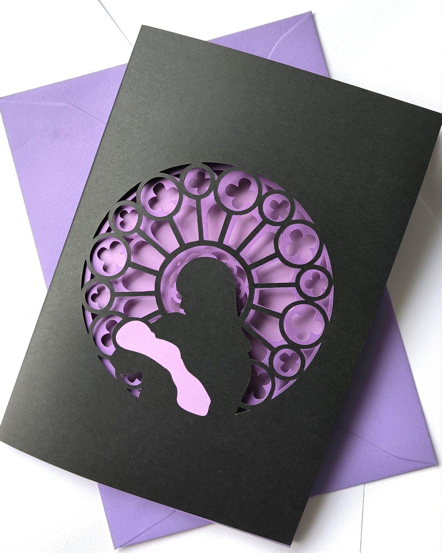 Handmade papercut greeting card, with a layered rose window in purple and black. Features a mother and baby silhouette in front of the gothic cathedral window design. Includes matching purple envelope.