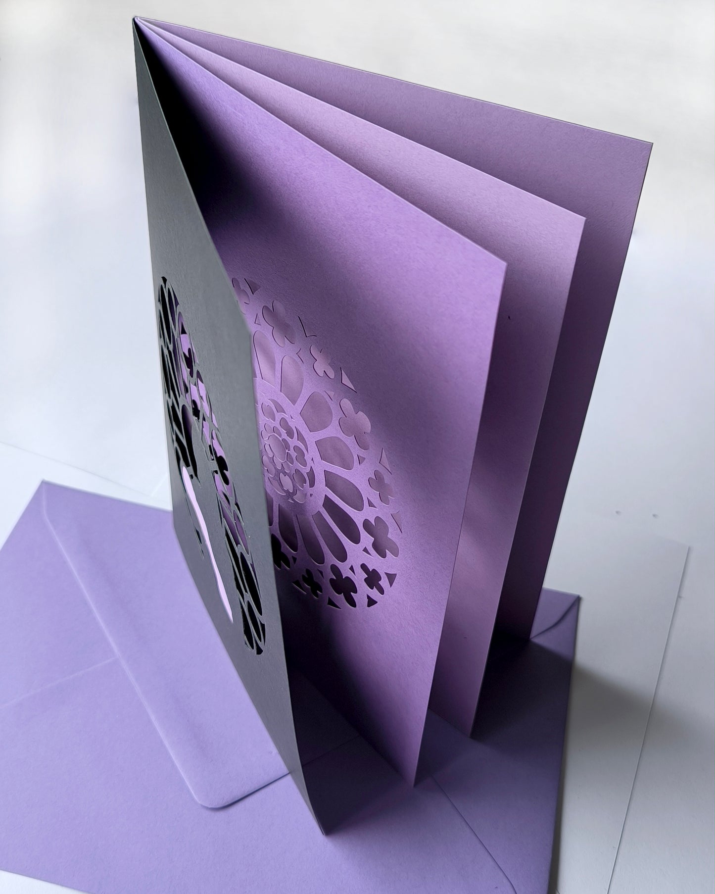 Handmade papercut greeting card, with a layered rose window in purple and black. Features a mother and baby silhouette in front of the gothic cathedral window design. Includes matching purple envelope.