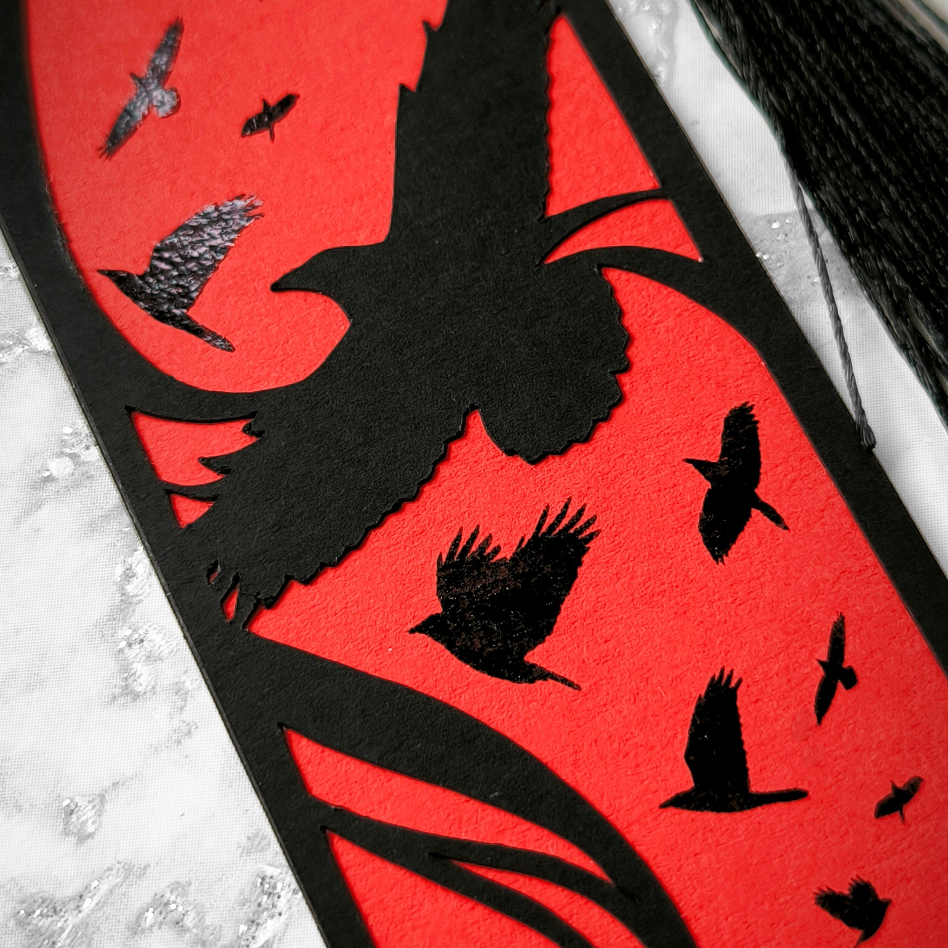 A handmade card bookmark. the black top layer is cut to a sweeping crow silhouette framed by flowing lines, set against a blood-red base scattered with glossy black-foiled crows in flight. Finished with a sleek black tassel.