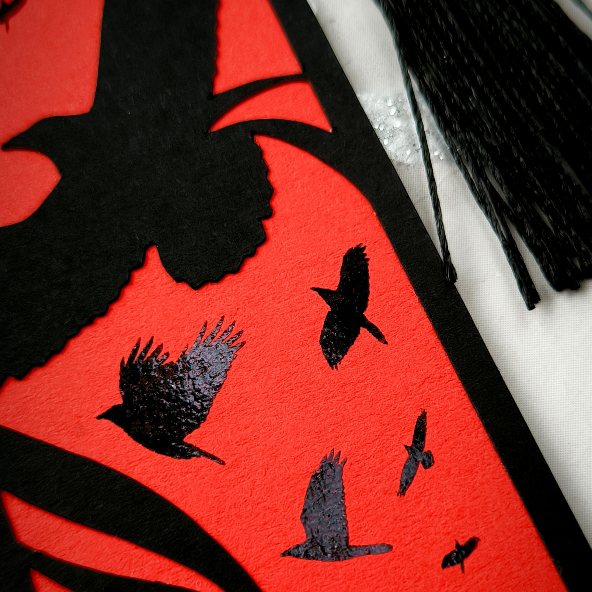A handmade card bookmark. the black top layer is cut to a sweeping crow silhouette framed by flowing lines, set against a blood-red base scattered with glossy black-foiled crows in flight. Finished with a sleek black tassel.