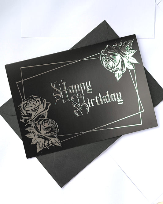 A smooth black card with raised silver chrome detailing. Silver gothic text reads Happy Birthday, surrounded by a silver rectangular frame and silver roses in two corners.