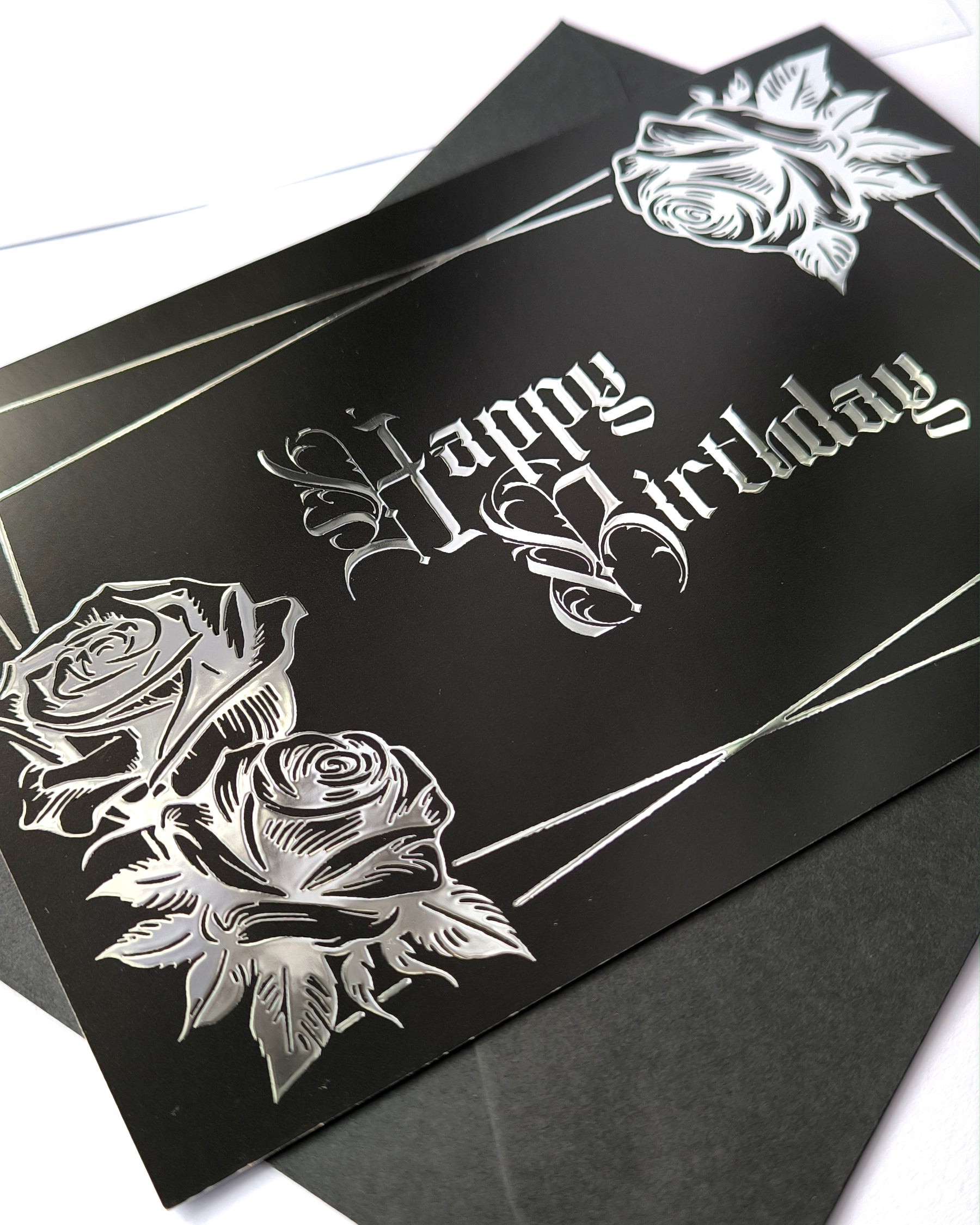 A smooth black card with raised silver chrome detailing. Silver gothic text reads Happy Birthday, surrounded by a silver rectangular frame and silver roses in two corners.