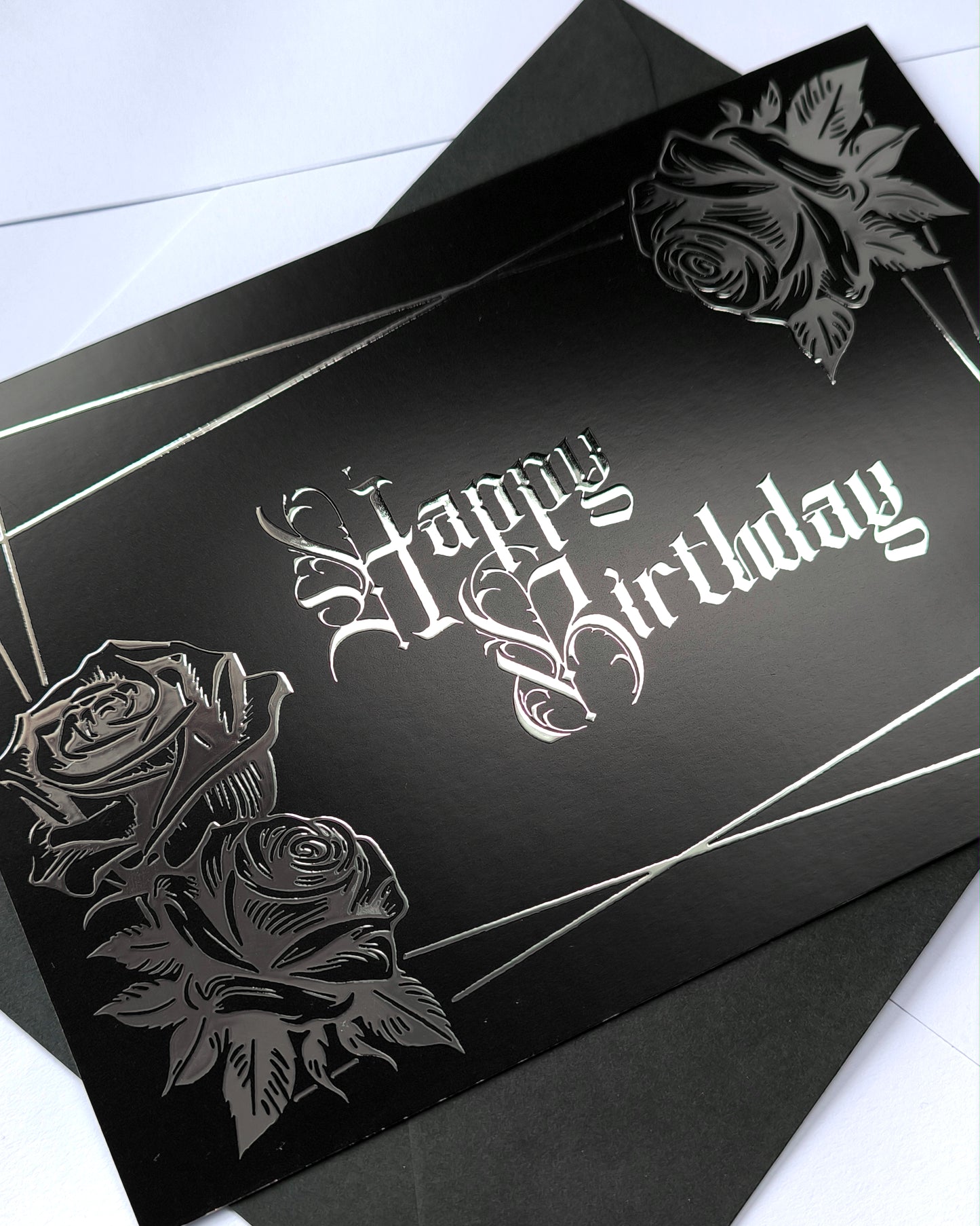 A smooth black card with raised silver chrome detailing. Silver gothic text reads Happy Birthday, surrounded by a silver rectangular frame and silver roses in two corners.