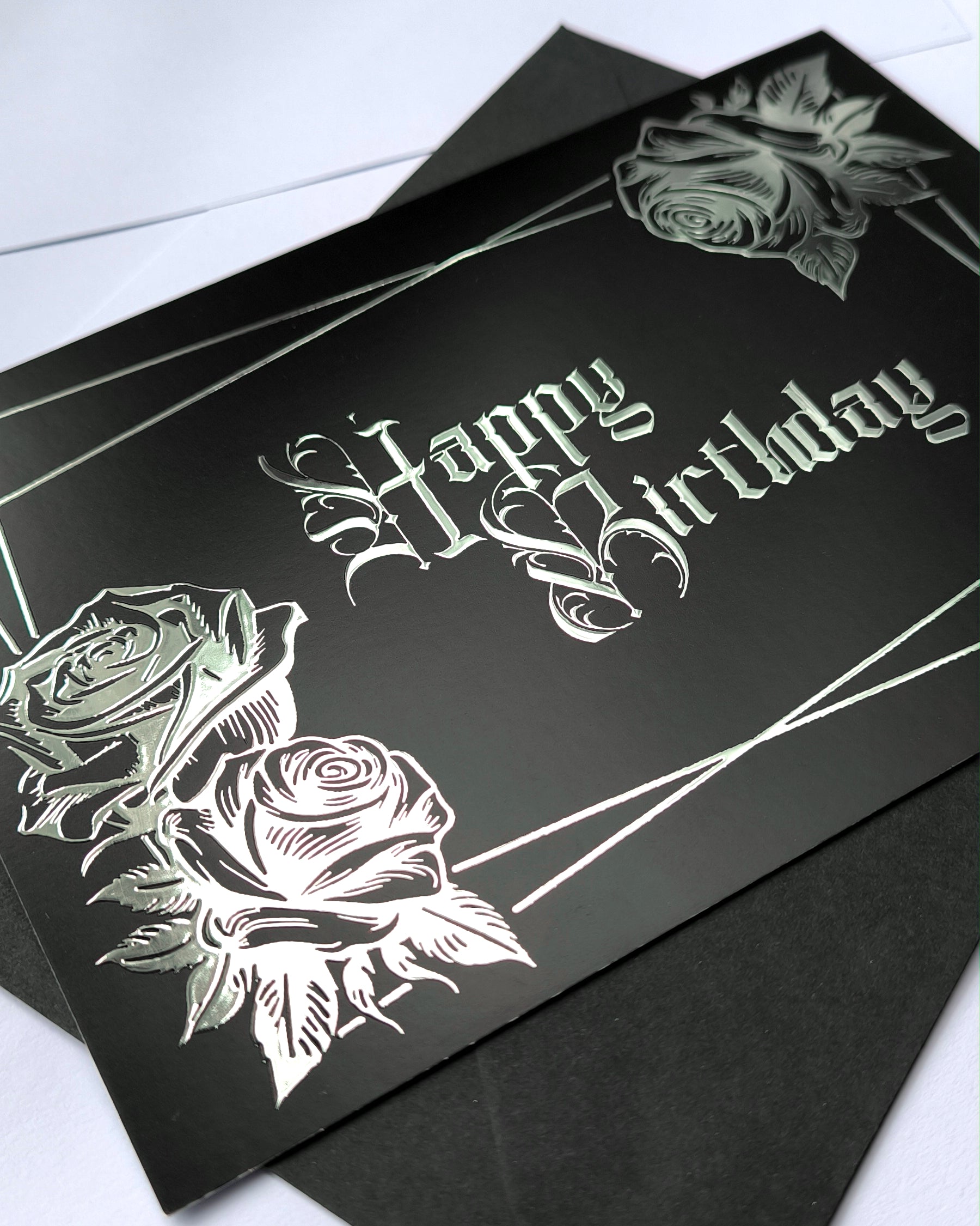 A smooth black card with raised silver chrome detailing. Silver gothic text reads Happy Birthday, surrounded by a silver rectangular frame and silver roses in two corners.