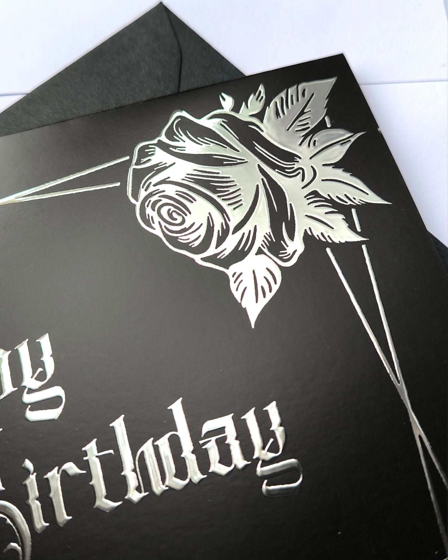 A smooth black card with raised silver chrome detailing. Silver gothic text reads Happy Birthday, surrounded by a silver rectangular frame and silver roses in two corners.