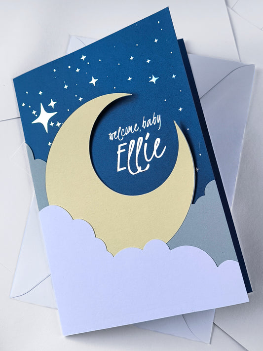 Handmade A5 layered papercut new baby card in deep navy with a cream crescent moon, white cloud layers, and silver foil star details, personalised with the phrase "welcome baby" and a custom name in silver foil script. The navy interior has a blank white cloud for your own message.