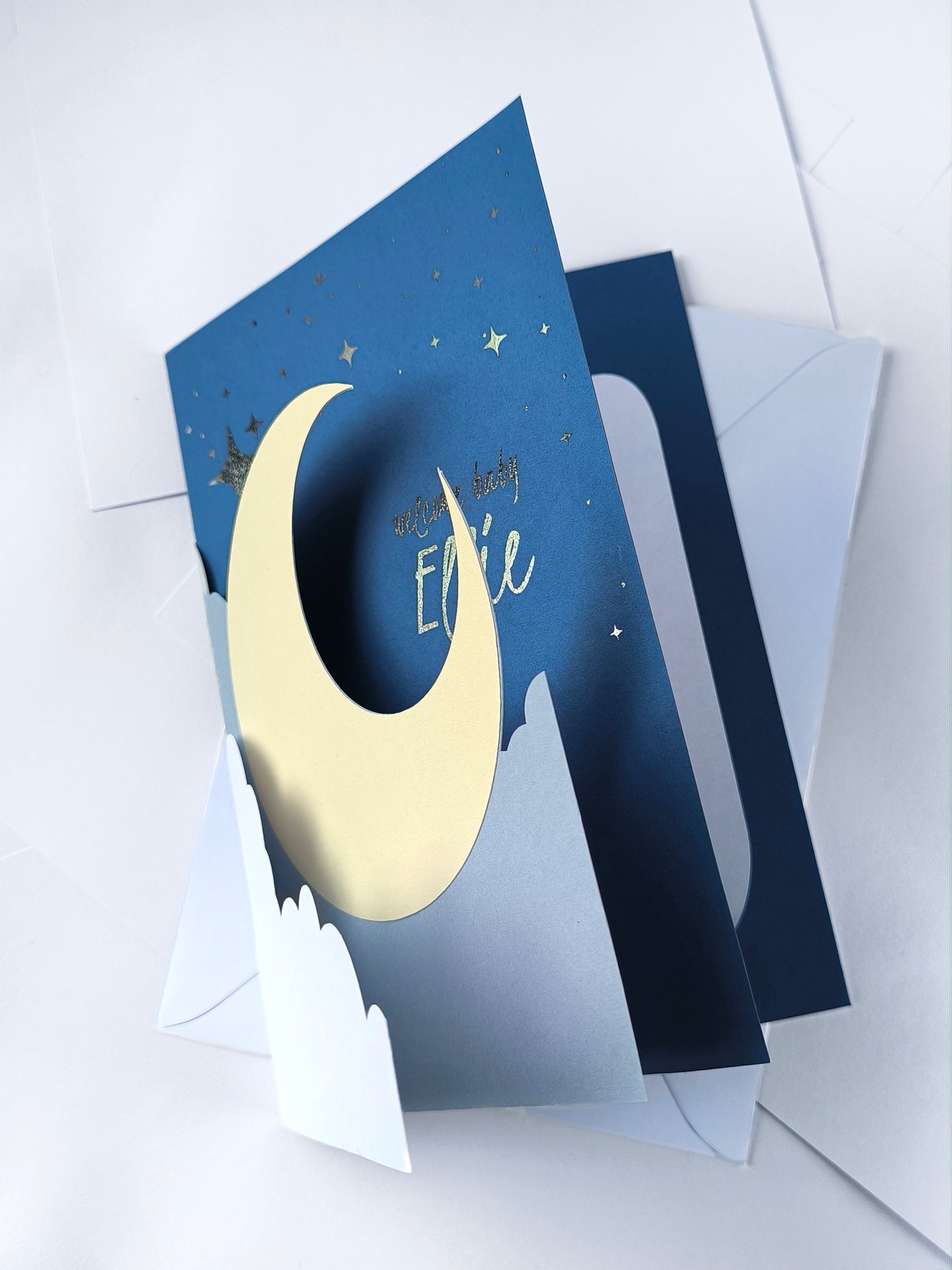Handmade A5 layered papercut new baby card in deep navy with a cream crescent moon, white cloud layers, and silver foil star details, personalised with the phrase "welcome baby" and a custom name in silver foil script. The navy interior has a blank white cloud for your own message.