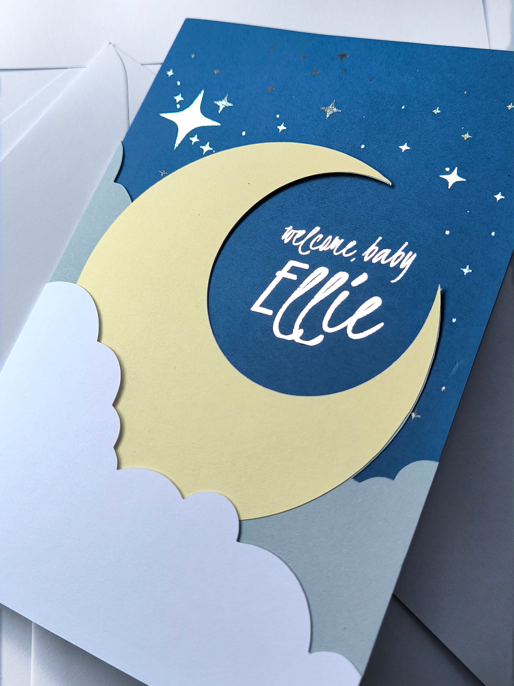 Handmade A5 layered papercut new baby card in deep navy with a cream crescent moon, white cloud layers, and silver foil star details, personalised with the phrase "welcome baby" and a custom name in silver foil script. The navy interior has a blank white cloud for your own message.