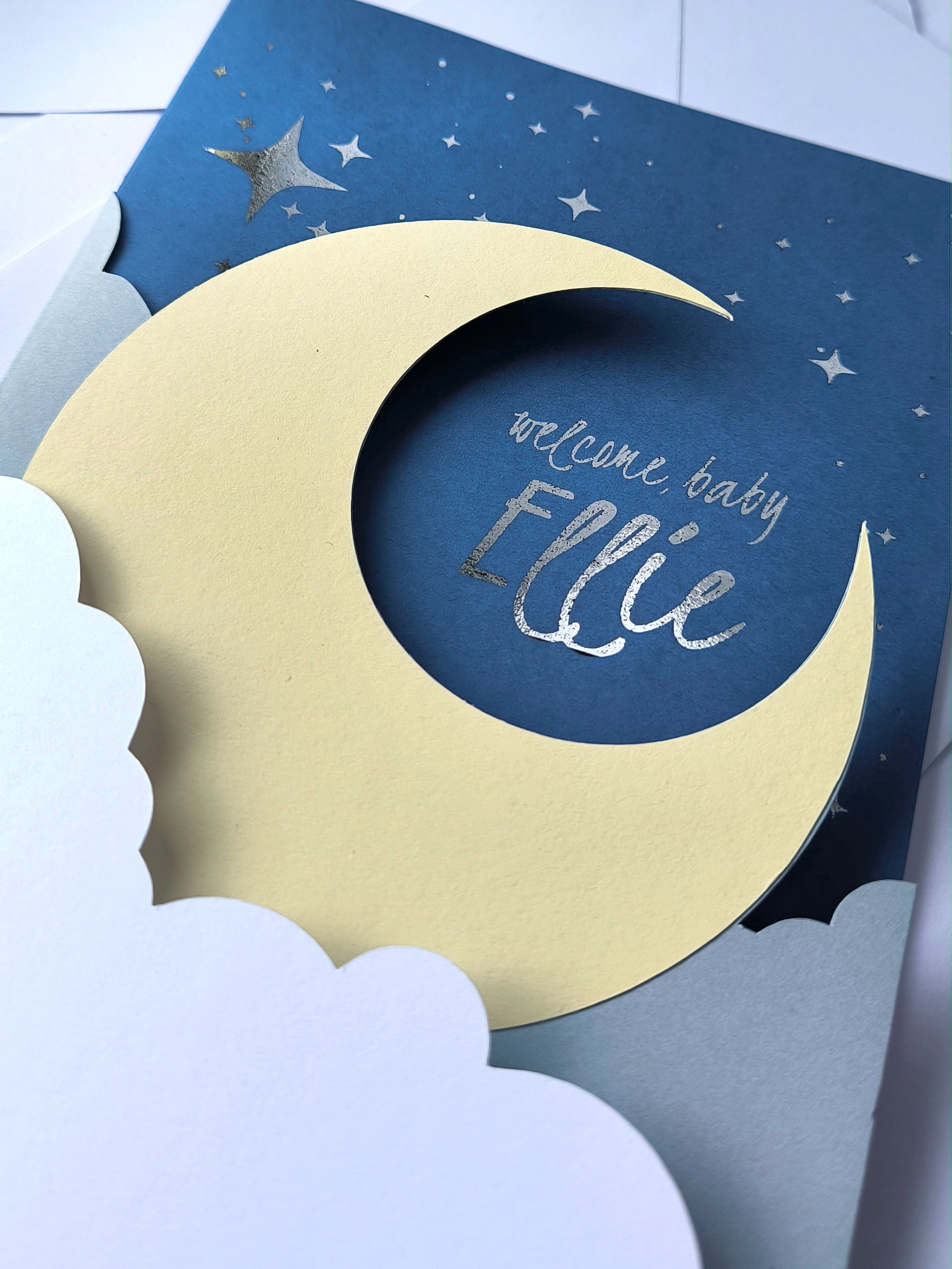 Handmade A5 layered papercut new baby card in deep navy with a cream crescent moon, white cloud layers, and silver foil star details, personalised with the phrase "welcome baby" and a custom name in silver foil script. The navy interior has a blank white cloud for your own message.