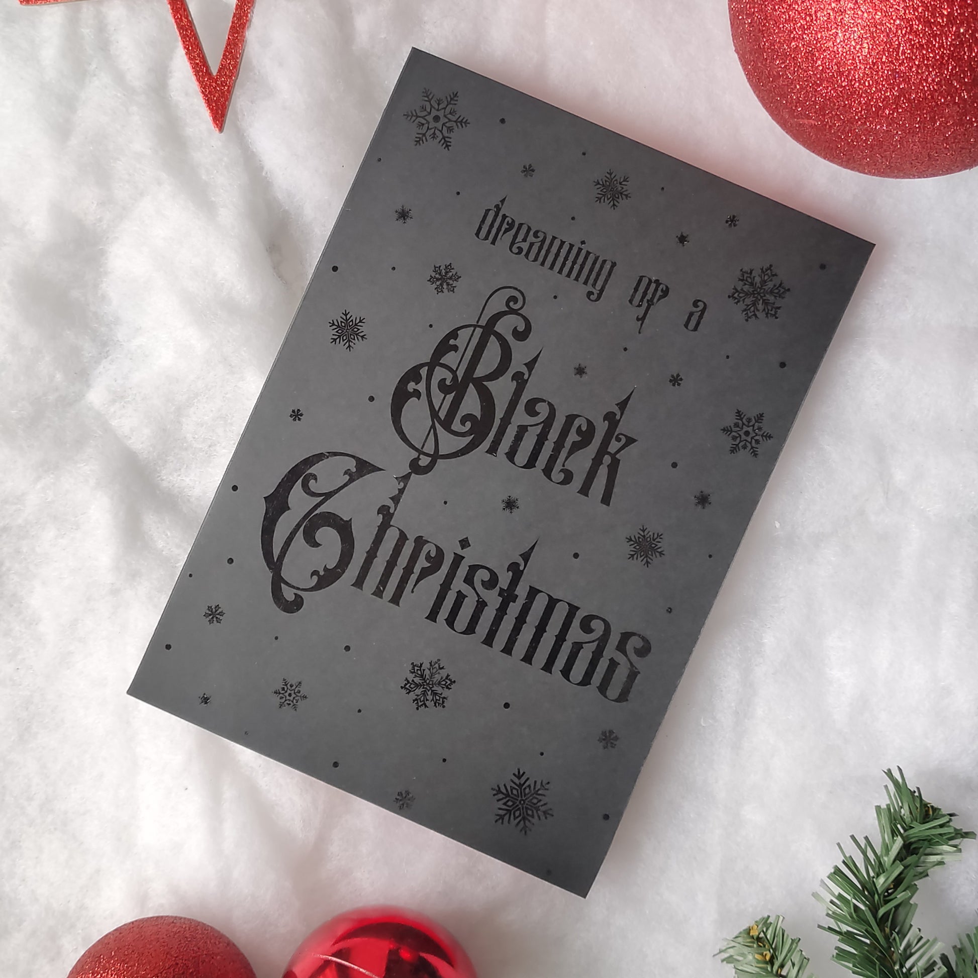 A5 handmade black Christmas card with black foil snowflakes and gothic lettering reading “Dreaming of a Black Christmas”. Portrait format on smooth black card with red interior. Includes red envelope. Blank inside.