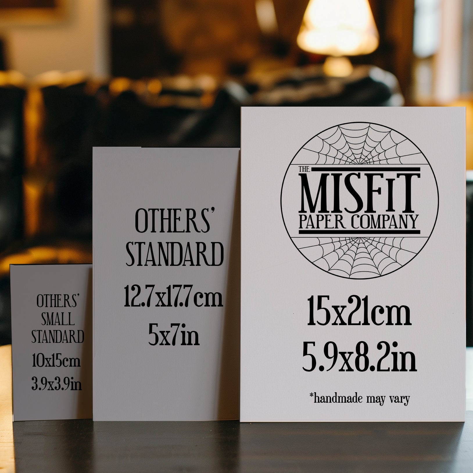 A size comparison image featuring three greeting cards standing upright on a table. The smallest card on the left is labeled "OTHERS’ SMALL STANDARD" (10x15cm / 3.9x3.9in). The middle card is labeled "OTHERS’ STANDARD" (12.7x17.7cm / 5x7in). The largest card on the right, branded with "The Misfit Paper Company" logo is labeled 15x21cm (5.9x8.2in) with a note that "handmade may vary." The image visually highlights that The Misfit Paper Co.’s cards are larger than typical standard sizes.