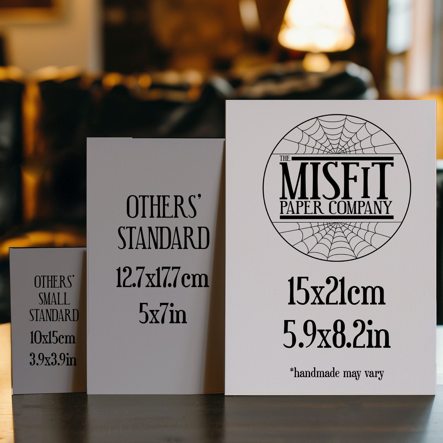 A size comparison image featuring three greeting cards standing upright on a table. The smallest card on the left is labeled "OTHERS’ SMALL STANDARD" (10x15cm / 3.9x3.9in). The middle card is labeled "OTHERS’ STANDARD" (12.7x17.7cm / 5x7in). The largest card on the right, branded with "The Misfit Paper Company" logo is labeled 15x21cm (5.9x8.2in) with a note that "handmade may vary." The image visually highlights that The Misfit Paper Co.’s cards are larger than typical standard sizes.