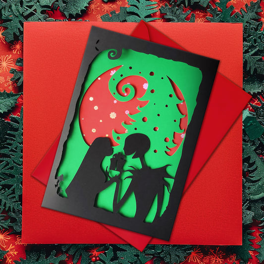 A5 handmade layered papercut Christmas card in black, red and green. Silhouette couple exchange a gift beneath a swirling tree and red moon with holographic snowflakes. Portrait format with depth. Includes red envelope. Blank inside.