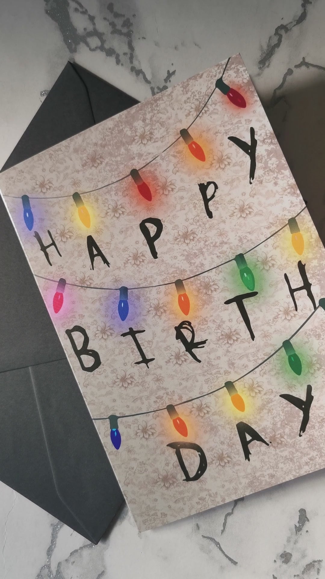 Stranger Things-inspired birthday card with fairy light lettering on a dark wall design, spelling “Happy Birthday” in a spooky, scrawled style over retro wallpaper.