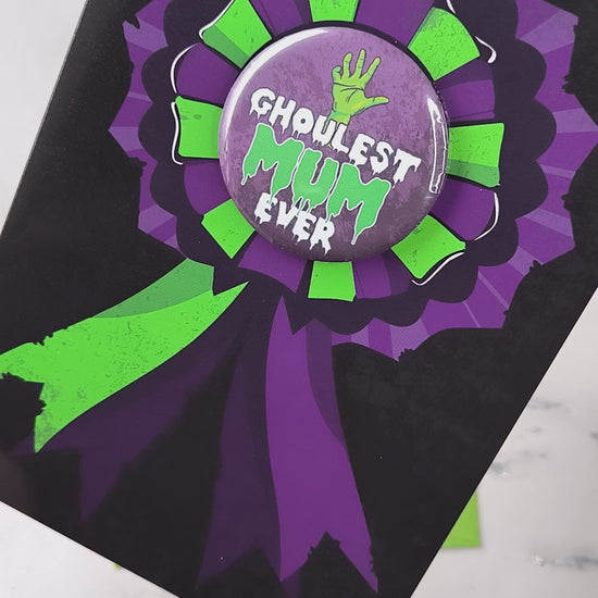 A large glossy A5 greeting card featuring a green and purple rosette with a removable 58mm badge reading "Ghoulest Mom Ever" in a bold, spooky font. Available in Mom, Mum, or Mam variations. The card is blank inside and comes with an envelope.