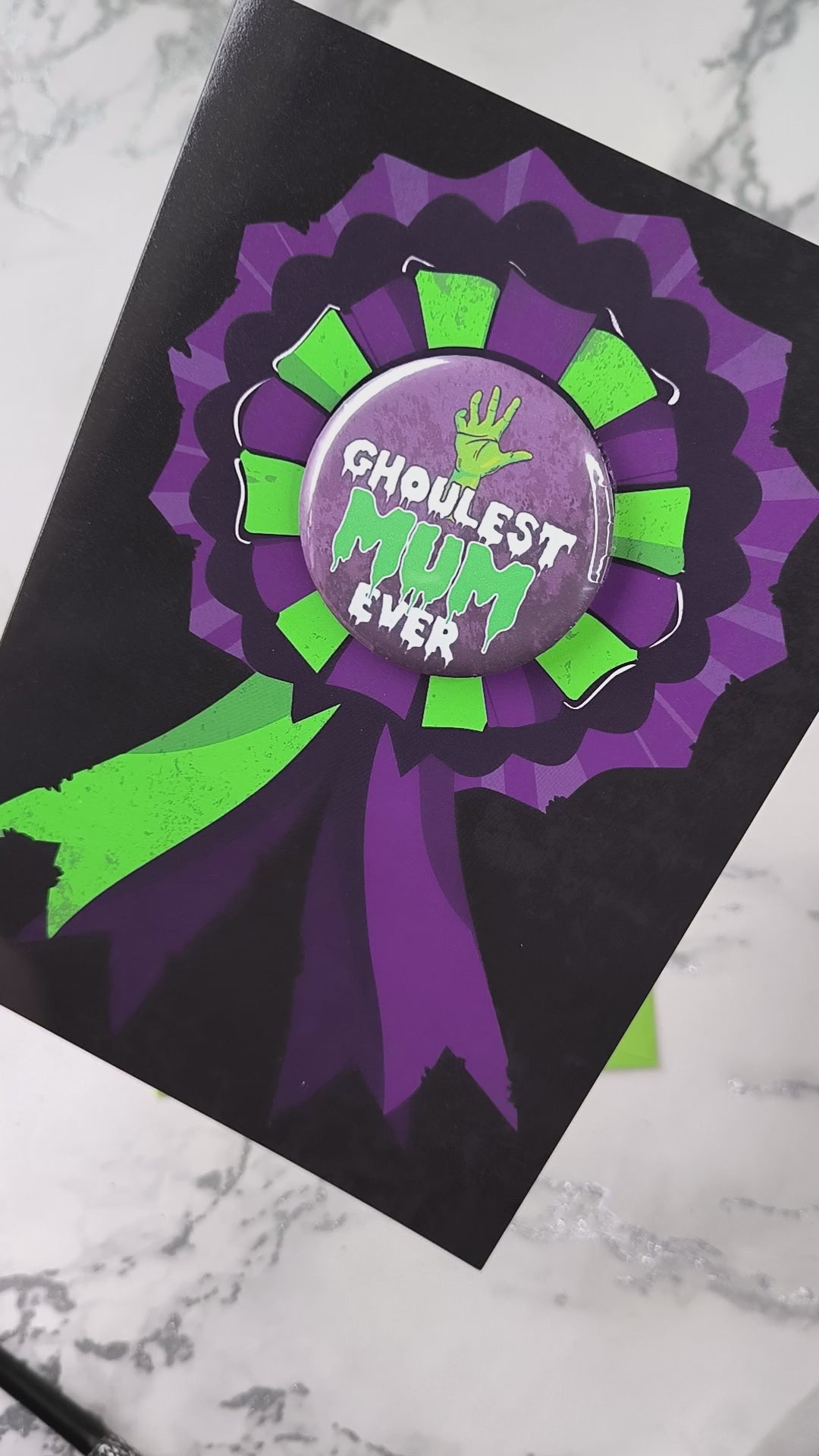 A large glossy A5 greeting card featuring a green and purple rosette with a removable 58mm badge reading "Ghoulest Mom Ever" in a bold, spooky font. Available in Mom, Mum, or Mam variations. The card is blank inside and comes with an envelope.