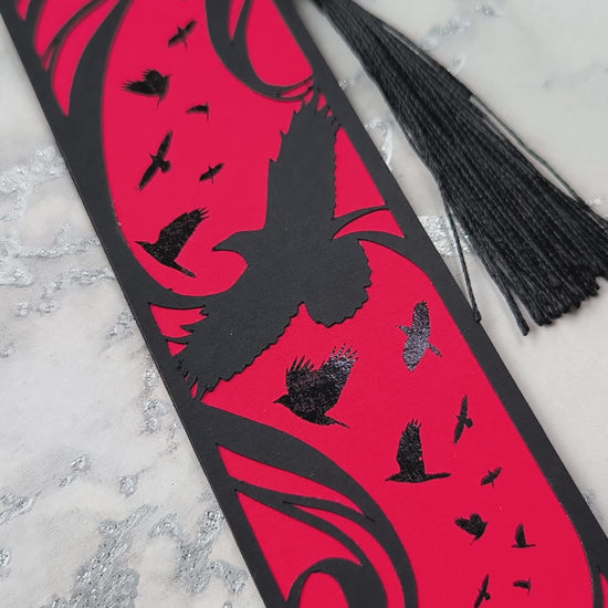 A handmade card bookmark. the black top layer is cut to a sweeping crow silhouette framed by flowing lines, set against a blood-red base scattered with glossy black-foiled crows in flight. Finished with a sleek black tassel.
