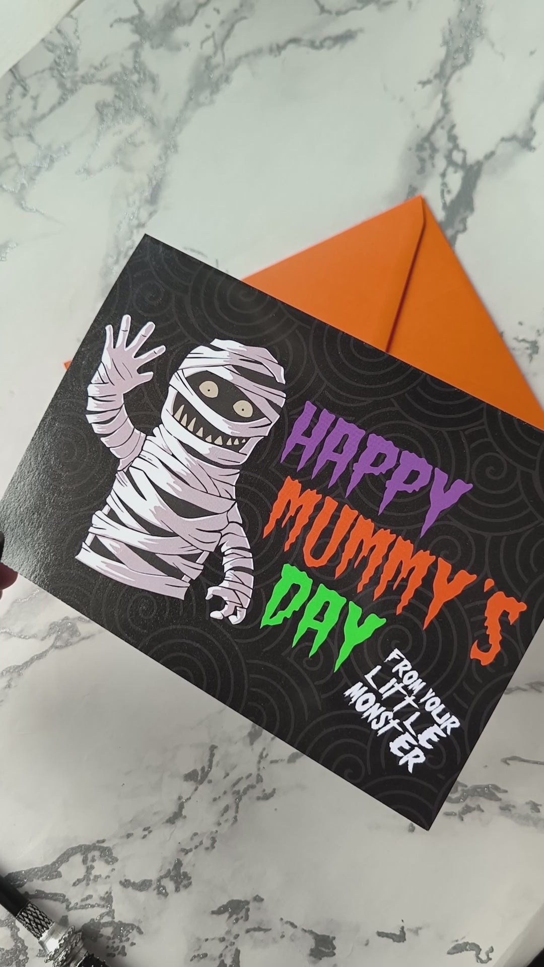A large landscape A5 greeting card featuring a spooky cute mummy illustration with the phrase "Happy Mummy’s Day" in bold Halloween colours. The inside is decorated with a spiral motif but has no text. Comes with a bright orange envelope.