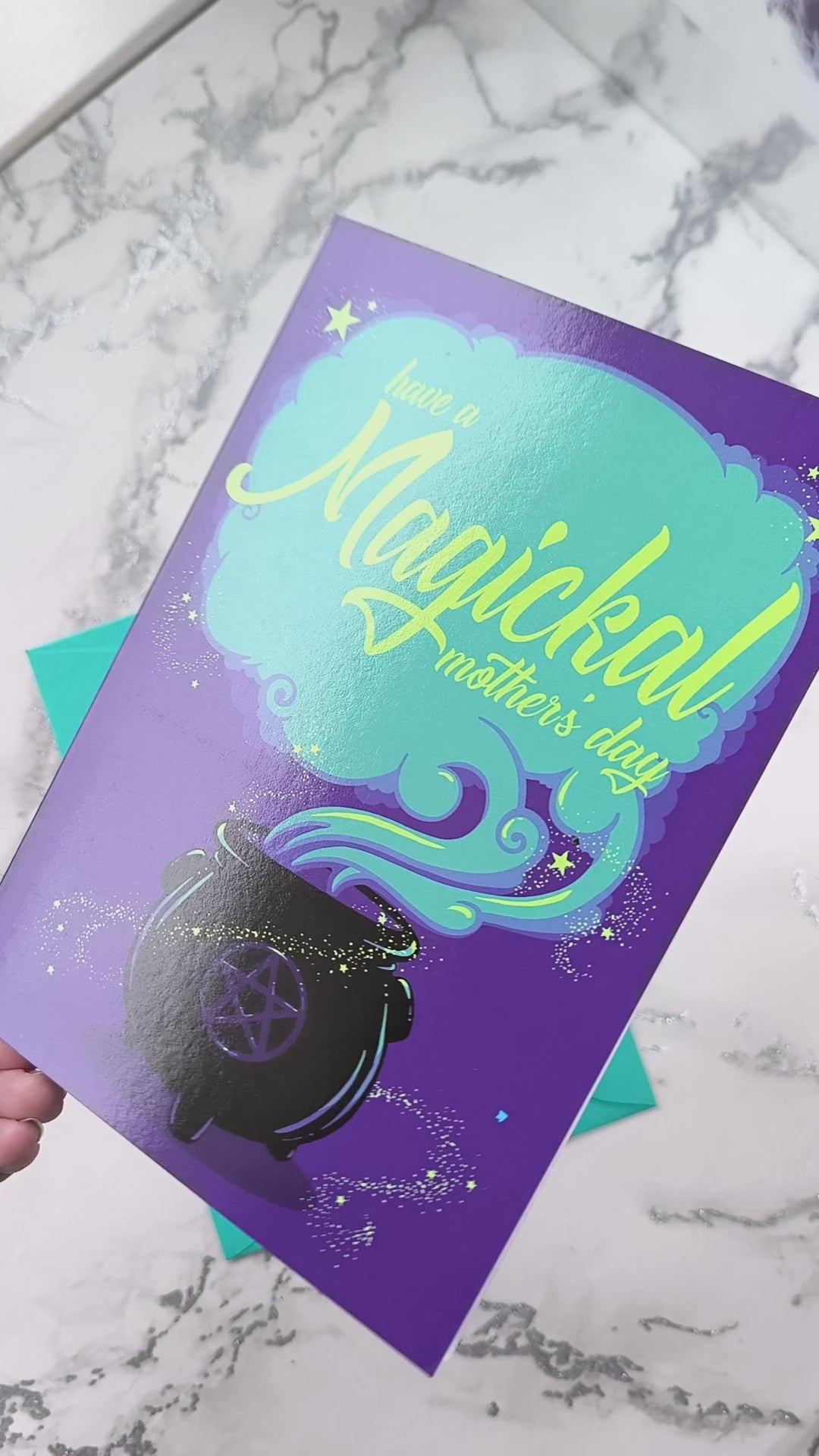 A large A5 greeting card in purple or pink, featuring a witchy cauldron illustration with billowing sparkling clouds and the words "Have a Magickal Mother’s Day." The inside has an illustration only (no text). Comes with an envelope included.
