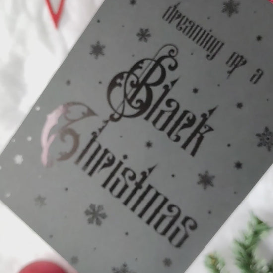 A5 handmade black Christmas card with black foil snowflakes and gothic lettering reading “Dreaming of a Black Christmas”. Portrait format on smooth black card with red interior. Includes red envelope. Blank inside.